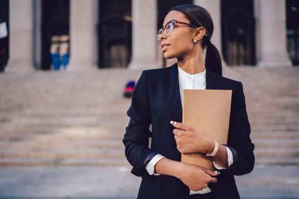 Pensive African American female lawyer in stylish formal suit holding folder with mock up area and looking away standing against courthouse. Half length of woman professional advocate with documents stock photo