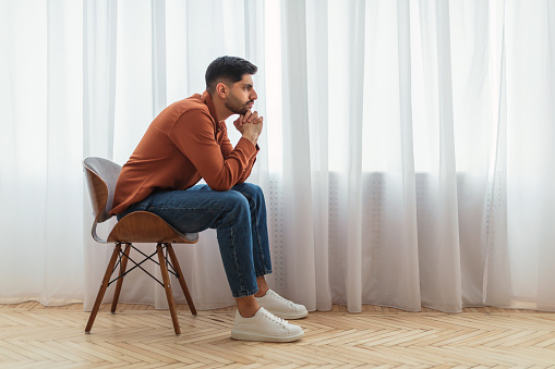 Pensive young man thinking sitting on chair at home in living room near window, profile side view portrait, free copy space. Thoughtful Arabic guy with serious face expression reflecting on problems