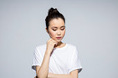 Portrait of worried asian young woman wearing white t-shirt, looking away with hand on chin. Studio shot, grey background.