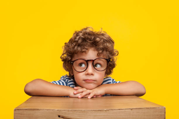 Pensive charming boy on studio background Portrait of curly boy in glasses leaning on box looking away pensively on orange background. eyewear stock pictures, royalty-free photos & images
