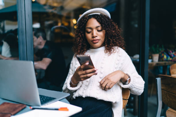 Pensive dark skinned millennial hipster girl watching videos browsed web page on smartphone, beautiful thinkign african american woman freelancer using banking app for paying and transaction online stock photo
