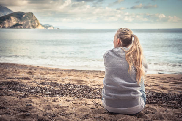 Pensive lonely young woman sitting on beach hugging her knees and looking into the distance with hope stock photo