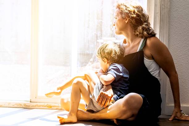 Pensive Mature woman posing with her son, sitting on the floor, very concerned looking through window worried about loss of her job and eviction due Covid-19 pandemic stock photo
