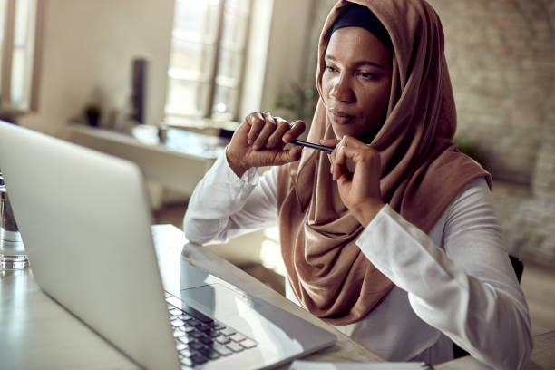 Pensive Muslim businesswoman working on laptop at her office desk. Black Islamic businesswoman reading an e-mail on a computer while working in the office. hijab stock pictures, royalty-free photos & images