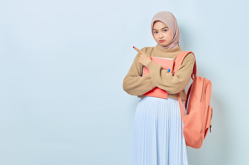 Pensive young Asian Muslim woman student in brown sweater with backpack, pointing fingers aside at copy space isolated on white background. back to school concept