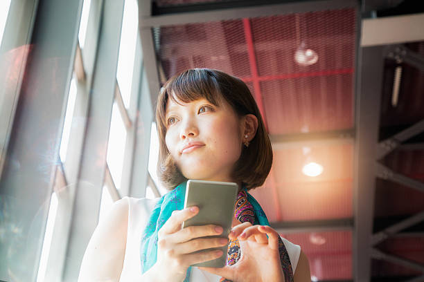 Pensive Young Japanese Woman daydreaming phone in hand Pensive Young Japanese Woman daydreaming phone in hand. Low angle view with an industrial ceiling in the background. shy japanese woman stock pictures, royalty-free photos & images