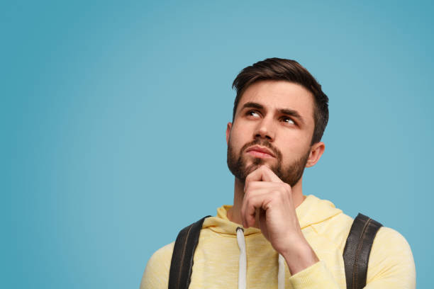 Pensive young man with backpack Handsome bearded guy with backpack looking up in doubts on studies against blue background hand on chin stock pictures, royalty-free photos & images