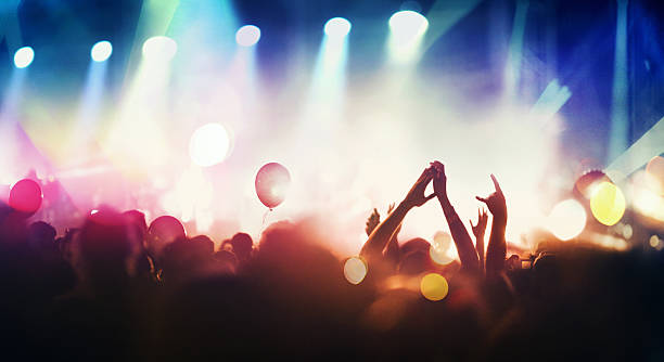 People at concert party. Rear view of large group of unrecognizable people enjoying a concert. Applauding, cheering, shooting video clips. One person in the crowd is holding a beer can. Band is performing on the stage, stage is toned purle and blue while the crowd is lit orange. staging light stock pictures, royalty-free photos & images