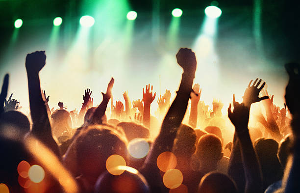 People at concert party. Rear view of large group of unrecognizable people enjoying a concert. Applauding, cheering, shooting video clips. Band is performing on the stage, stage is toned purple and blue while the crowd is lit orange. staging light stock pictures, royalty-free photos & images
