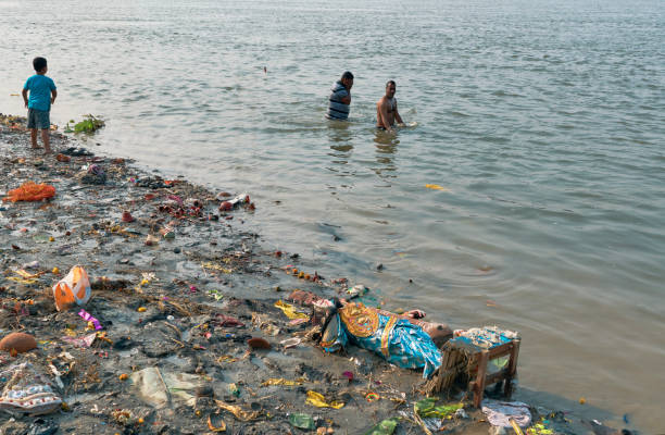 People bathing in Ganges, near a heavily polluted ghat Kolkata, 02-02-2020: Heavily polluted embankment of Ganges at Judges Ghat, with piled garbages and structures of clay idols dumped in water, after immersion of Goddess Saraswati idols in to Ganges. Two people are seen bathing in background. ganges river stock pictures, royalty-free photos & images