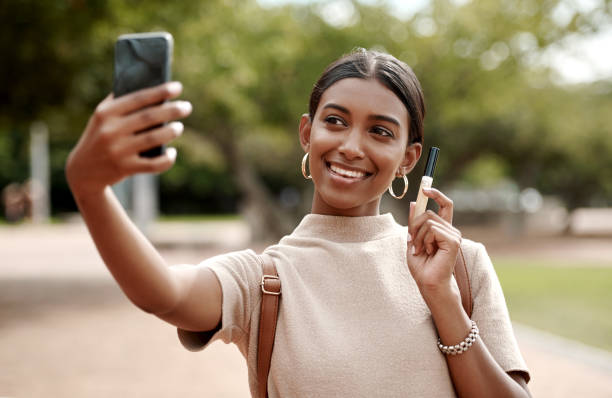 People connect to the person more than the product Shot of a young businesswoman taking selfies with a smartphone and holding a tube of lipgloss  against a city background influencer stock pictures, royalty-free photos & images