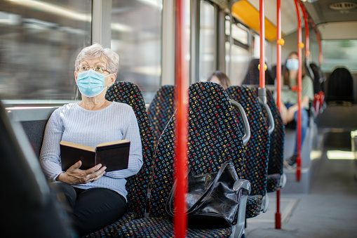 People in public bus transport. Senior woman sitting and reading a book. She wears protective face mask. Other people are in the bus
