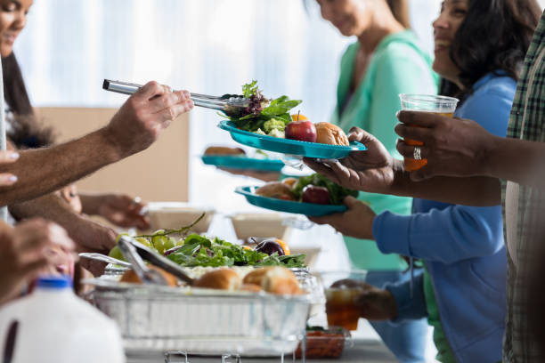 People receive food from volunteers in soup kitchen Volunteers serve healthy meals to people in soup kitchen or homeless shelter. Salad, fruit and rolls are served to the people in line. community dinner stock pictures, royalty-free photos & images