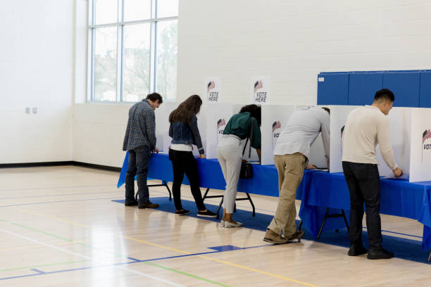 People stand at voting booths along the gym wall People of varying ages stand and vote at the voting booths set up against the wall of the school gym. voting photos stock pictures, royalty-free photos & images