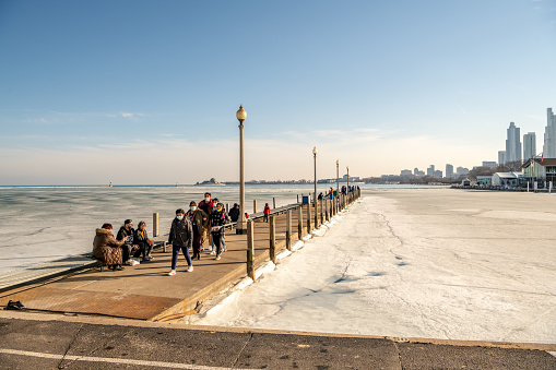 Chicago, IL, USA - February 27,2021: People walk along a pier on Lake Michigan, off the Lakefront Trail downtown, in winter, with the lake frozen over beneath and the skyline beyond.