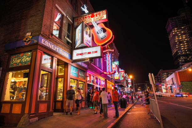 People walk along Broadway in Nashville Tennessee USA at night Nashville, Tennessee - June 20, 2019:  People walk by the famous Ernest Tubb Record Shop by the businesses and bars along Broadway in Nashville Tennessee USA.  Broadway is known for its honky-tonks, Music and record stores. broadway nashville stock pictures, royalty-free photos & images