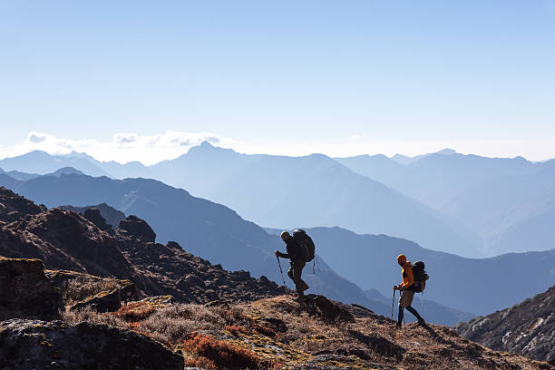 People with Backpacks and trekking Sticks traveling in Mountains Two Men walking on rocky slope carrying Backpacks using trekking Sticks dressed in alpine Jackets and hiking Pants. Layered Mountains View beside Silhouetted of People mountain ridge stock pictures, royalty-free photos & images