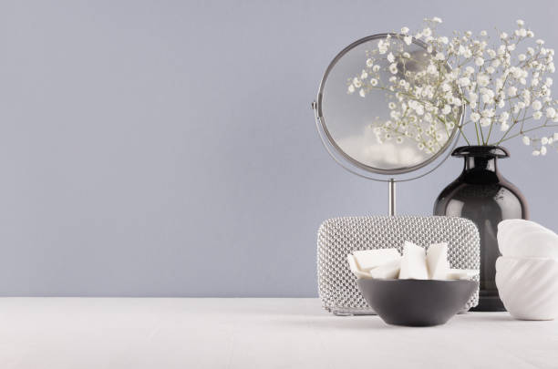 Photo of Perfect stylish decoration for home in grey colors - black glass vase with small fluffy flowers, mirror, female silver cosmetic bag, bowl sponges on soft ligth white wood table.
