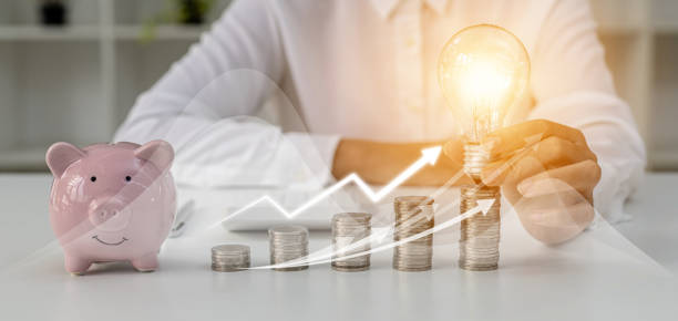 A person holds a light bulb on top of a pile of coins, a large number of coins are placed on the table from low to high, financial chart graphics and growth arrows. personal finance concept. stock photo