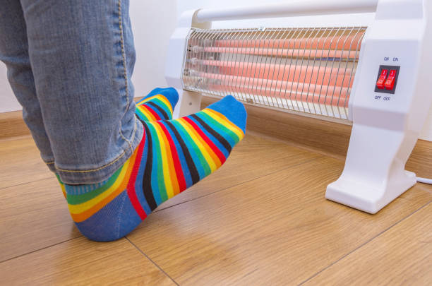 eine person, die helle regenbogenfarbene socken trägt und kalte füße in der nähe einer elektrischen heizung wärmt. infrarot-halogenheizung zu hause. - heizung stock-fotos und bilder