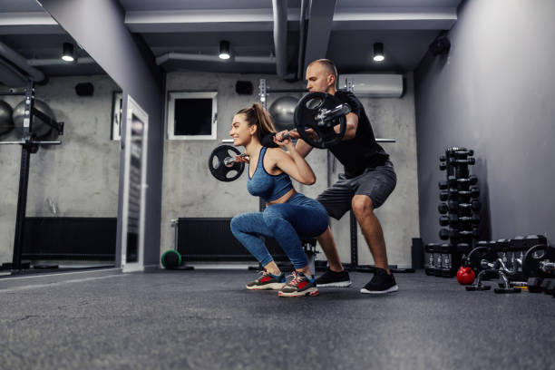 Personal training in the gym. A young woman and in sportswear and in good shape, she does barbell squats to strengthen the muscles of the whole body. The assistance of trainers in individual training stock photo
