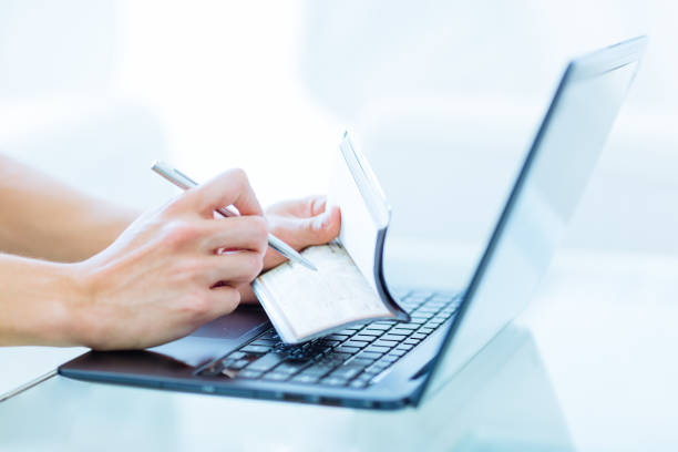 Persons hands writing a personal bank cheque using a pen while working on a laptop computer. Closeup of a caucasian male adult writing on an empty personal chequebook while using a laptop computer in a modern environment. bank account stock pictures, royalty-free photos & images