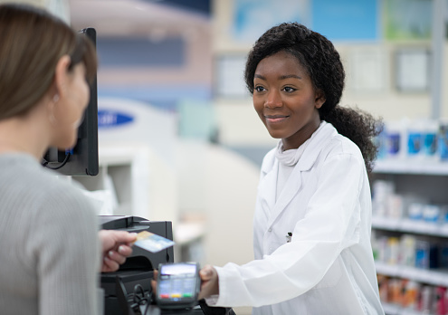A female pharmacist of African decent is seen standing behind the counter as she rings in a customers prescription purchase and checks her merchandise out.  She is wearing a white lab coat and has a friendly smile on her face.