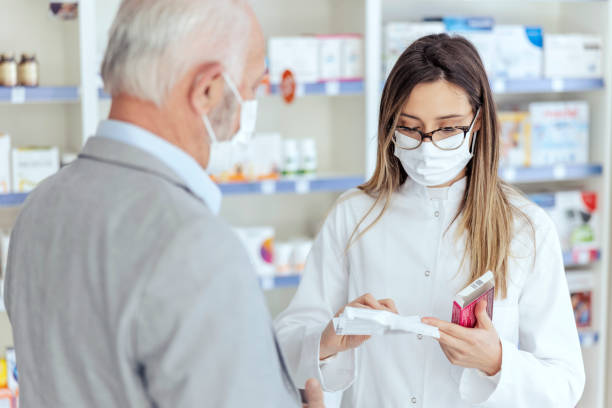 Pharmacist help Man asks for advice from a pharmacist regarding therapy prescribed by a doctor. A female pharmacist with glasses reads the declaration from the medicine They both wear protective masks stock photo