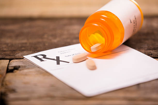 Pharmacy medication bottle with prescription pad. Pills spilling out. White, blank doctor's prescription pad with an orange prescription medication bottle lying on top of it with pills spilling out of the bottle.  The bottle has a blank label on it.  Tan colored pills lay on top of prescription.  A large "RX" is at the top left corner of the page. Pharmacy, healthcare, drugs, medicine concepts.  Desk, table, countertop. generic drug stock pictures, royalty-free photos & images