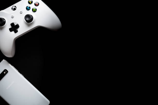 A phone and controller sit side by side as gaming becomes multi-platform Shot taken from above against a dark wooden background showing a White Samsung Galaxy S10+ and a Xbox One White Controller next to each other with room for text on the right hand side xbox stock pictures, royalty-free photos & images