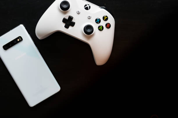 A phone and controller sit side by side as gaming becomes multi-platform Shot taken from above against a dark wooden background showing a White Samsung Galaxy S10+ and a Xbox One White Controller next to each other with room for text on the right hand side xbox photos stock pictures, royalty-free photos & images