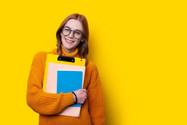 Photo of a caucasian redhead woman holding copybooks isolated on yellow color background with copyspace. Smiling face. stock photo