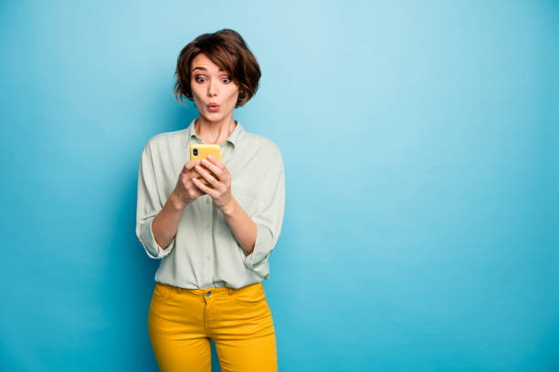 魅力的な女性の写真は、新しいブログ記事のコメントを読んで電話の手を持って、目はカジュアルな緑のシャツ黄色のズボンを着用し、青い色の背景を分離 - 驚く ストックフォトと画像