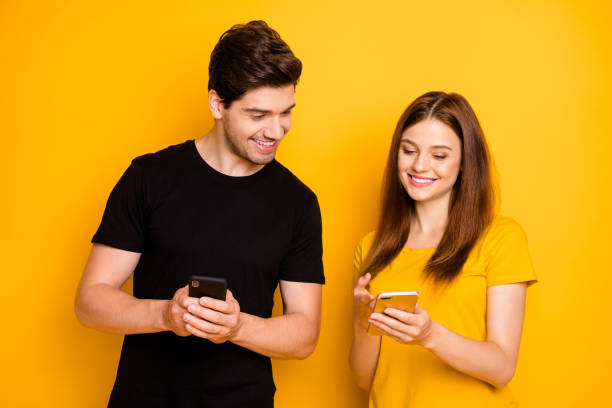 photo of cheerful positive cute charming nice influencers sharing expirience between each other in black yellow t-shirt holding telephones searching new information isolated vivid color background - friends color background imagens e fotografias de stock