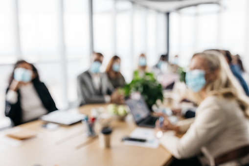 A blurred photo of a business meeting in a bright office. Smartly-dressed co-workers are wearing protective face masks in order to prevent the spread of COVID-19 virus. Their faces are not recognizable. Horizontal daylight indoor photo.