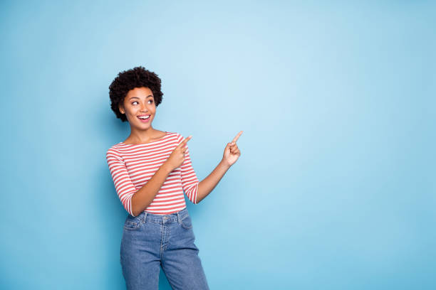 Photo of curly wavy cheerful positive nice pretty cute girlfriend pointing into empty space smiling toothily in jeans denim striped t-shirt isolated over blue pastel color background Photo of curly wavy cheerful positive nice pretty cute girlfriend pointing into empty space smiling toothily in jeans denim striped t-shirt, isolated over blue pastel color background pointing stock pictures, royalty-free photos & images