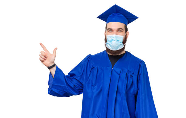 Photo of student man in bachelor clothes and graduating cap pointing away while wearing facial mask Photo of student man in bachelor clothes and graduating cap pointing away while wearing facial mask. medical degrees online stock pictures, royalty-free photos & images