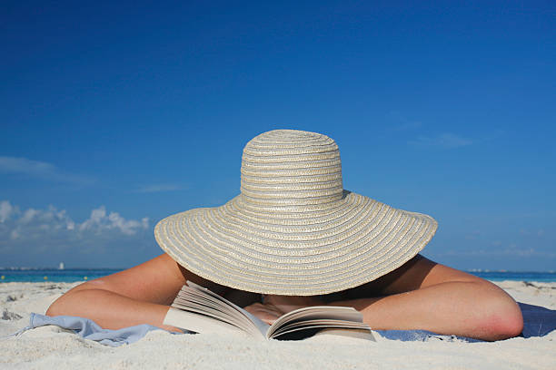 Photograph of woman lying on beach wearing sun hat reading stock photo