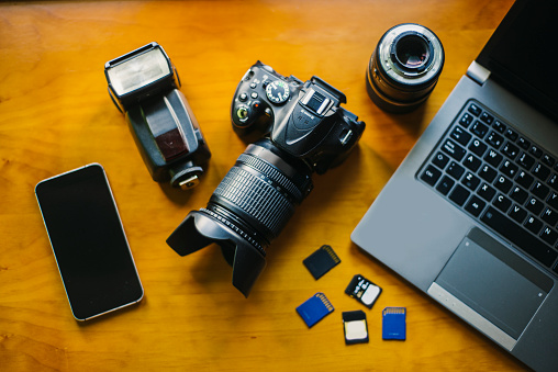 The desk of a photographer, including toold as the camera, lenses, flashes, SD cards, a mobile phone and a computer.