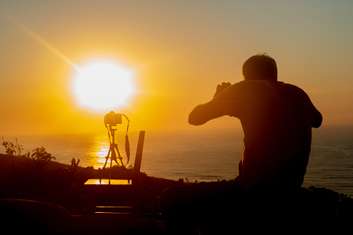 A silhouette of a man taking photographs with a camera in-front of an orange sun reflected in the ocean.