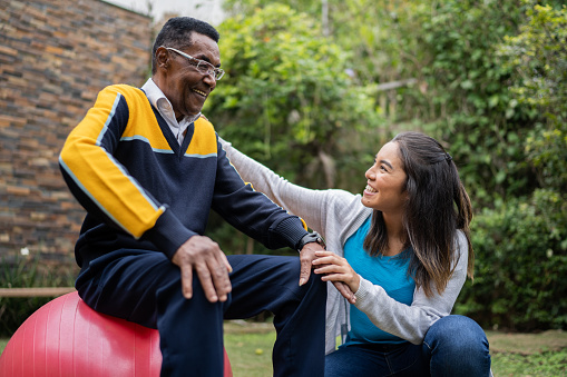 Physical therapist talking to senior man sitting on a fitness ball at home