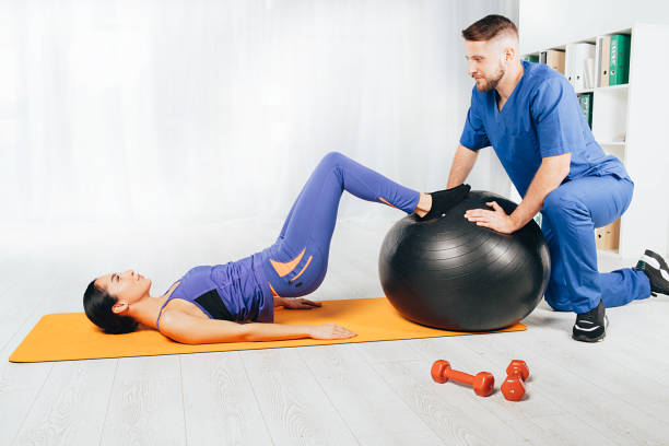Physical therapist working with a young woman. She lying on exercise mat and doing exercise with pilates ball Physical therapist working with a young woman. She lying on exercise mat and doing exercise with pilates ball yoga ball work stock pictures, royalty-free photos & images