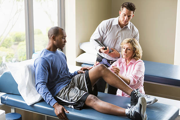 Physical therapists examining patient Physical therapists examining a young African American man sitting on a treatment table.  The female PT is using  a goniometer to measure the range of motion of his knee, while her male assistant watches. orthopedics stock pictures, royalty-free photos & images