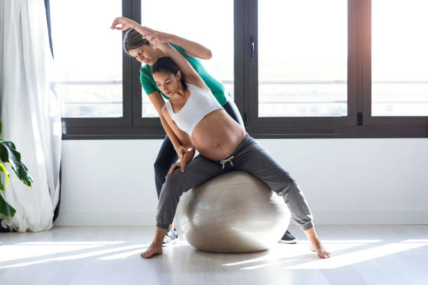 Physiotherapist helping to beautiful pregnant woman for doing pilates exercises with ball preparing for childbirth. stock photo