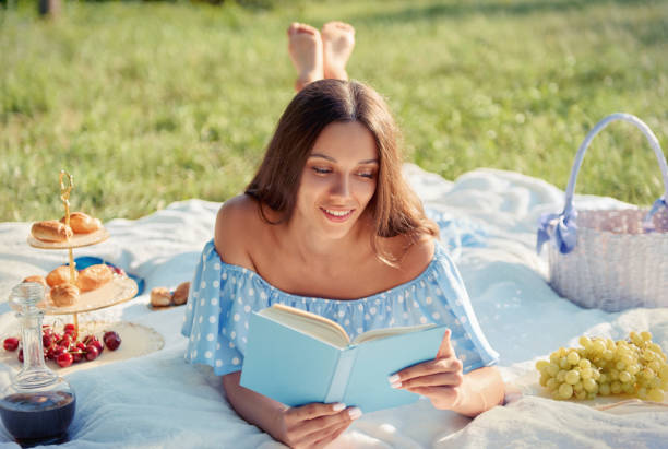 Picnic scene: pretty smiling girl lying on plaid and reading book in garden. Outdoor portrait of beautiful joyful young woman in blue dress stock photo