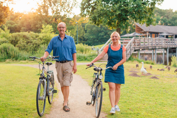 beeld van hoger paar dat met fietsen in het park bij zonsondergang loopt. - pensioen nederland stockfoto's en -beelden