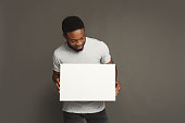 Picture of young smiling african-american man holding white blank board on grey background, copy space