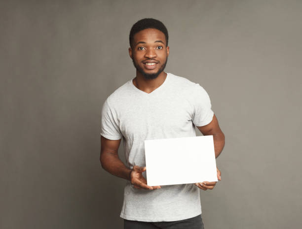 Picture of young african-american man holding white blank board Picture of young smiling african-american man holding white blank board for advertisement on grey background, copy space holding photos stock pictures, royalty-free photos & images