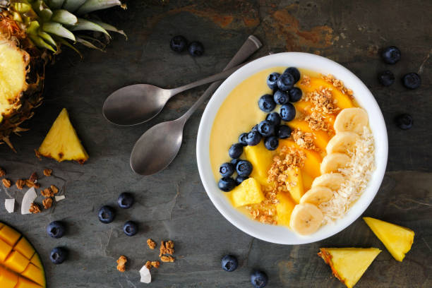 Pineapple, mango smoothie bowl with coconut, bananas, blueberries and granola, top view table scene on slate Healthy pineapple, mango smoothie bowl with coconut, bananas, blueberries and granola. Top view table scene on a dark background. mango smoothie stock pictures, royalty-free photos & images