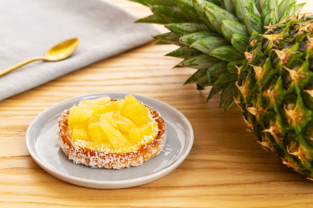 Pineapple tart or tartlet with grated coconut by an entire pineapple fruit and a grey napkin with a golden spoon, all on a wooden background. stock photo